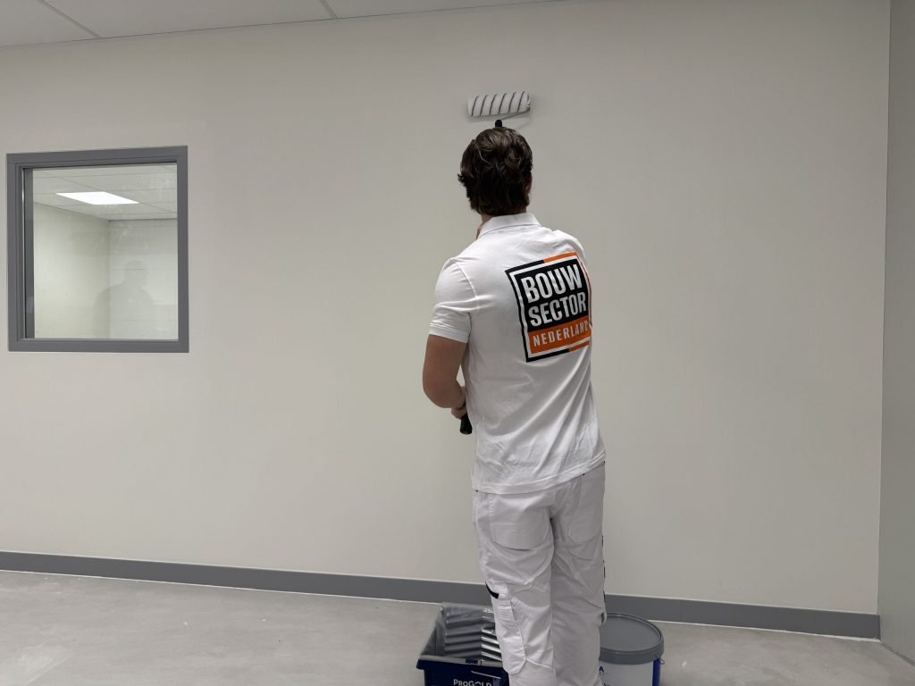 Schilder in wit werktenue zet de roller bovenaan een lichte wand, met verfbak en emmer op de vloer