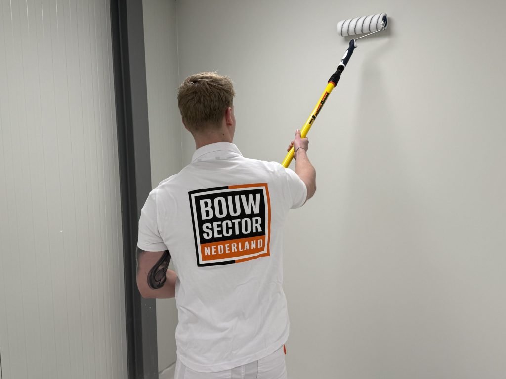 Schilder met roller aan een gele steel werkt een lichte wand bovenin af