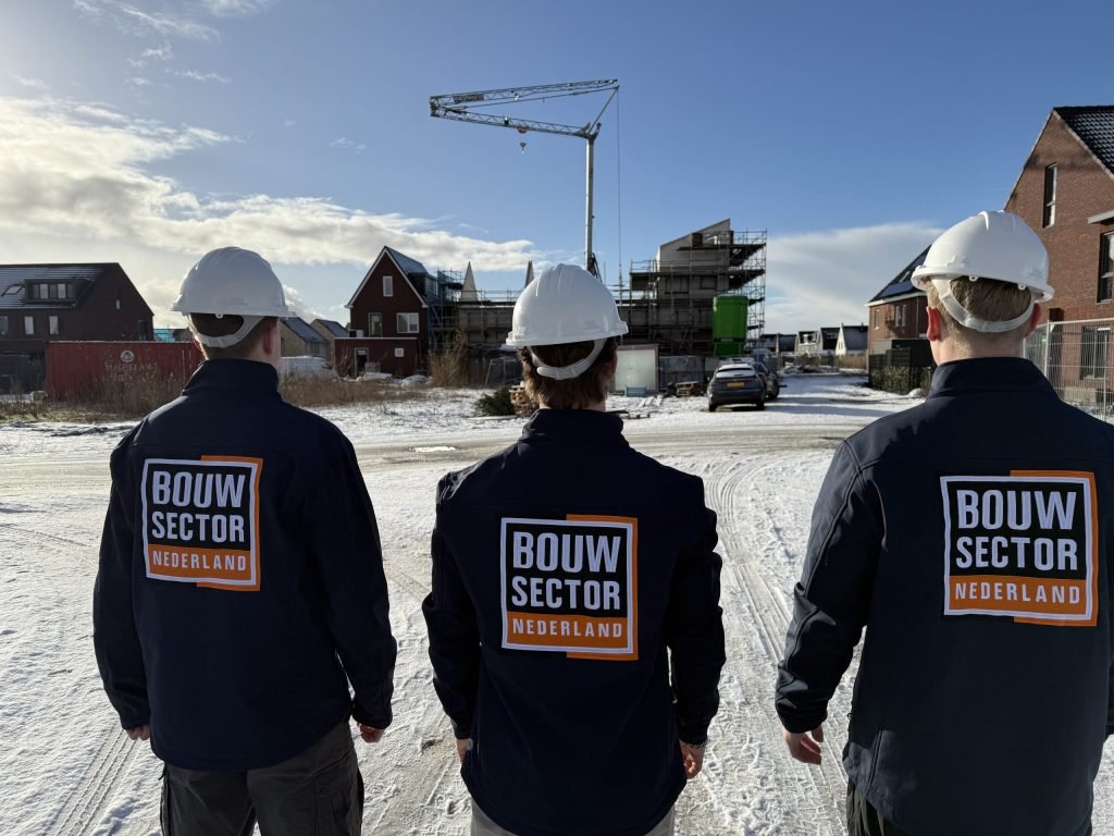 Drie vakmensen van Bouwsector Nederland met witte helm kijken uit over nieuwbouwwoningen in de sneeuw