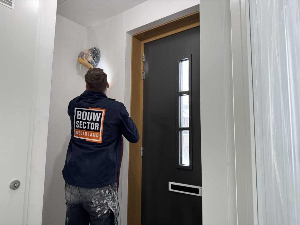 Vakman van Bouwsector Nederland schuurt met een wandschuurmachine de wand vlak onder het plafond in een hal