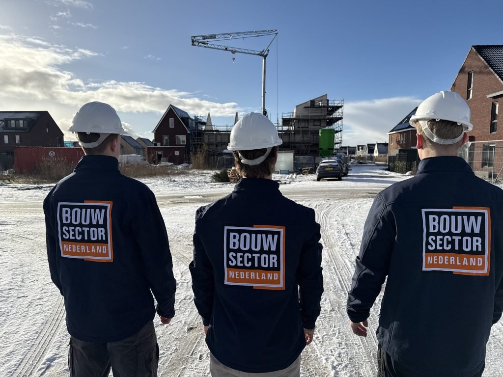 Drie vakmensen van Bouwsector Nederland met witte helm op een besneeuwde bouwweg bij nieuwbouwwoningen in aanbouw